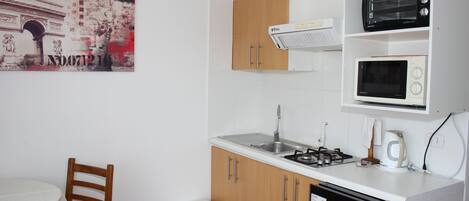 A compact kitchen with a sink, stove, and microwave. There is a small white stool and a wooden chair.