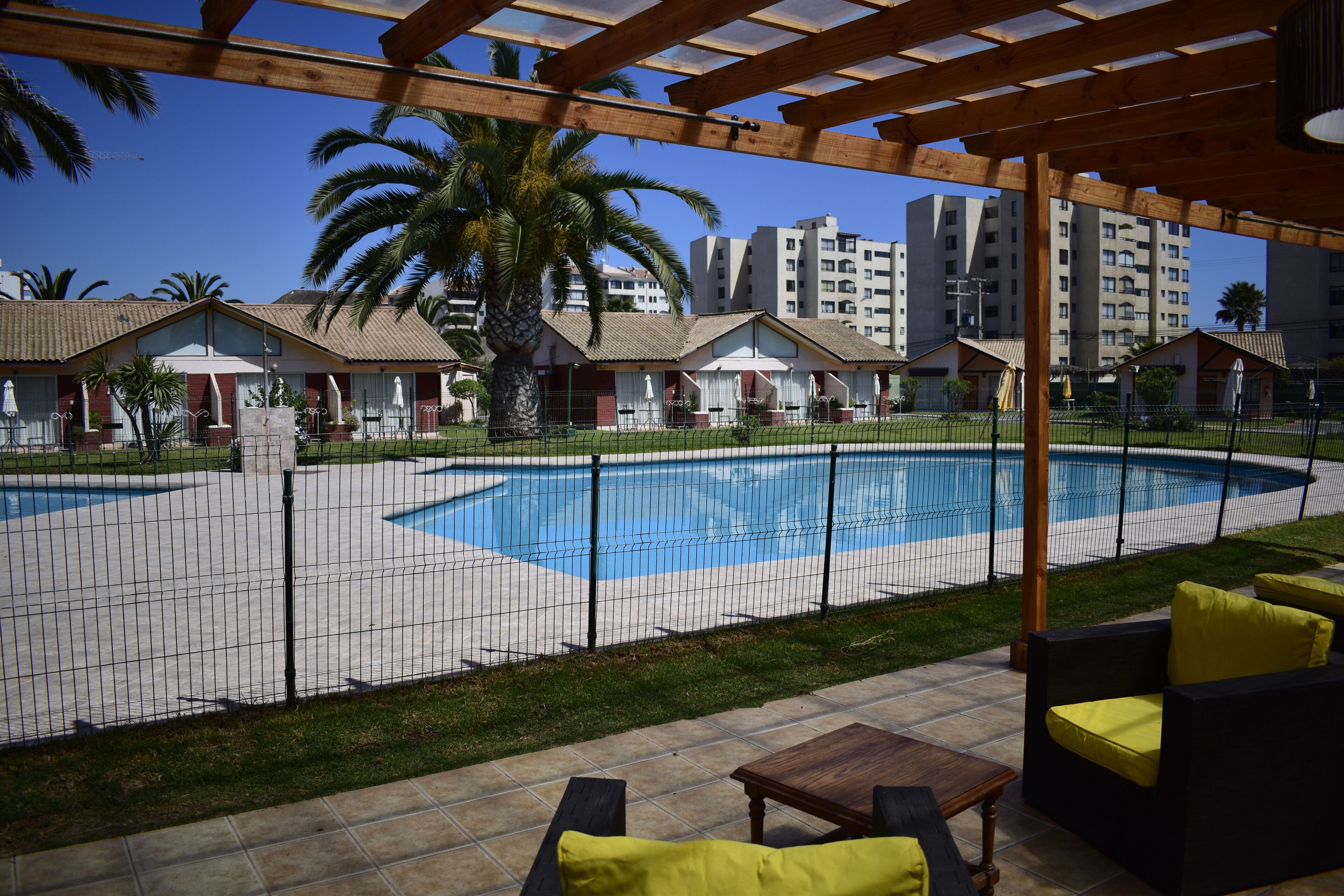 Ein Poolbereich mit einer Holzpergola, Sitzgelegenheiten im Freien und Blick auf Wohnhäuser im Hintergrund.