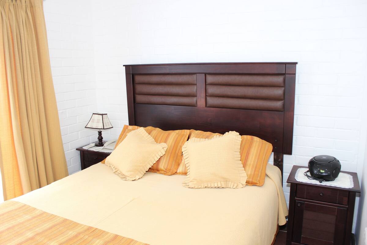 A neatly made bed with a headboard, a bedside lamp, and a small bedside table with a device on it.