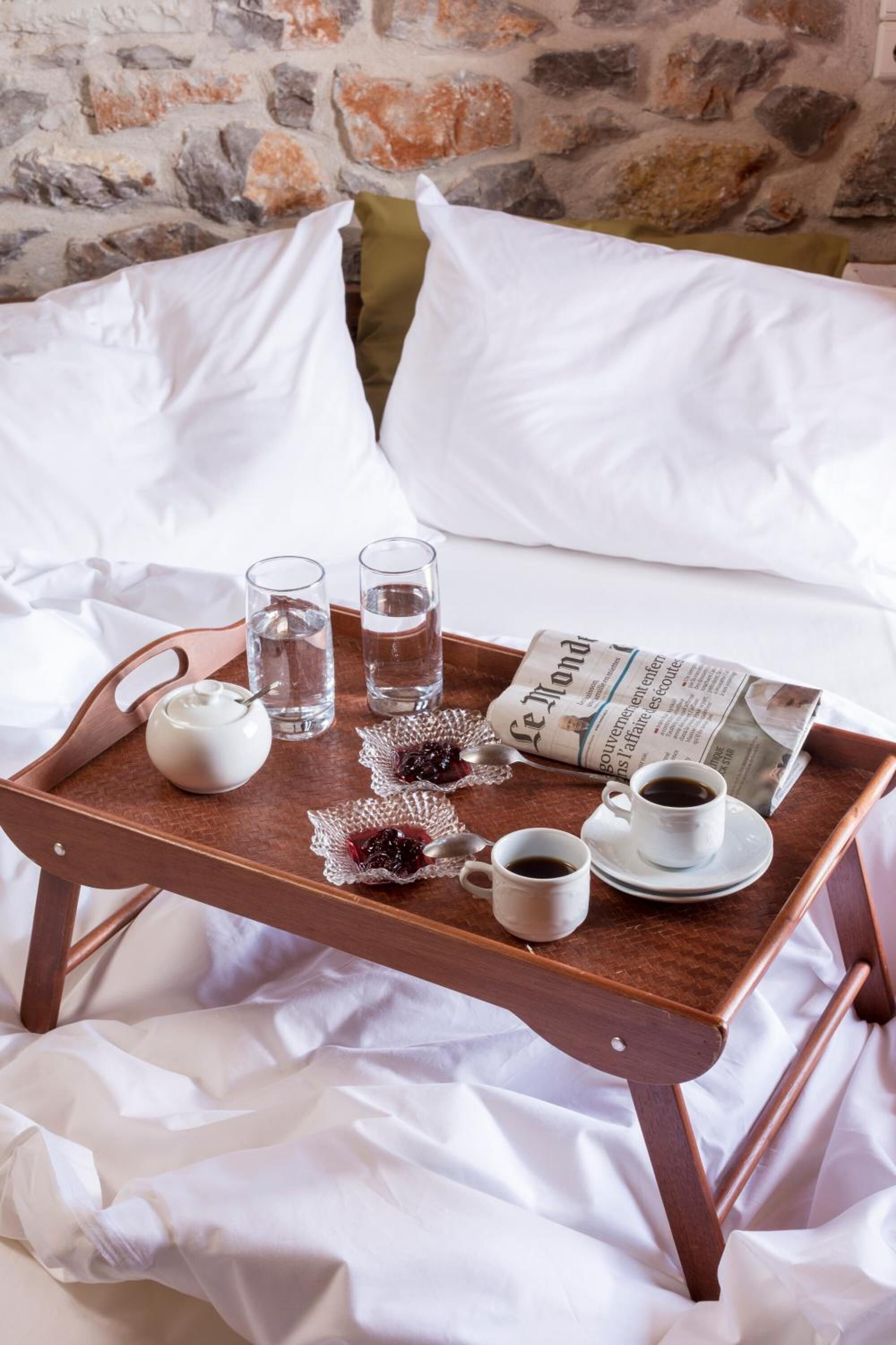 A wooden tray with a newspaper, a teapot, glasses of water, and cups of coffee on a bed with white sheets and pillows.