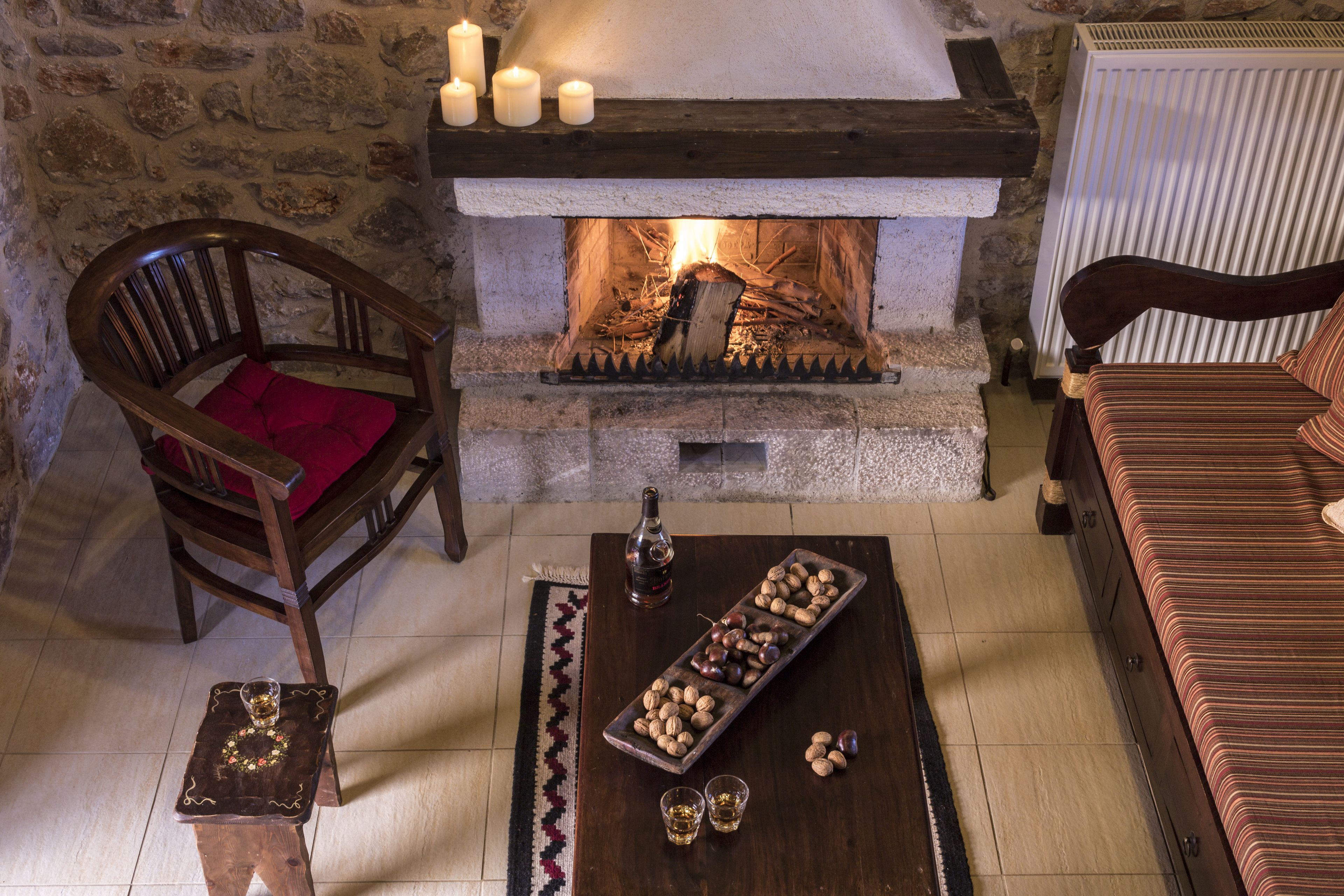 A cozy room with a stone wall, a fireplace with lit candles, a wooden chair with a red cushion, a wooden table with a tray of nuts and two shot glasses, and a striped sofa.
