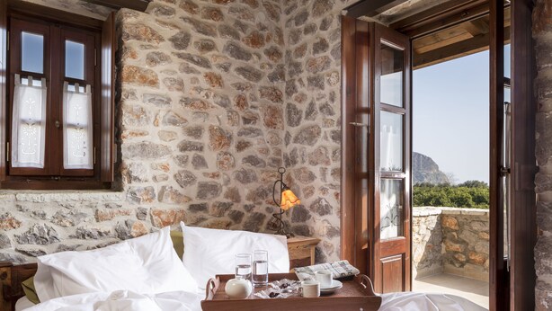 A bedroom with stone walls, a wooden bed, a tray with glasses and a teapot, and a view of a mountain. - Agroktima (South Kynouria)