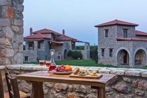 A stone table with a fruit platter, cheese, and glasses of red wine, set against a stone wall with a view of a stone building with red-tiled roofs. - Agroktima (South Kynouria)
