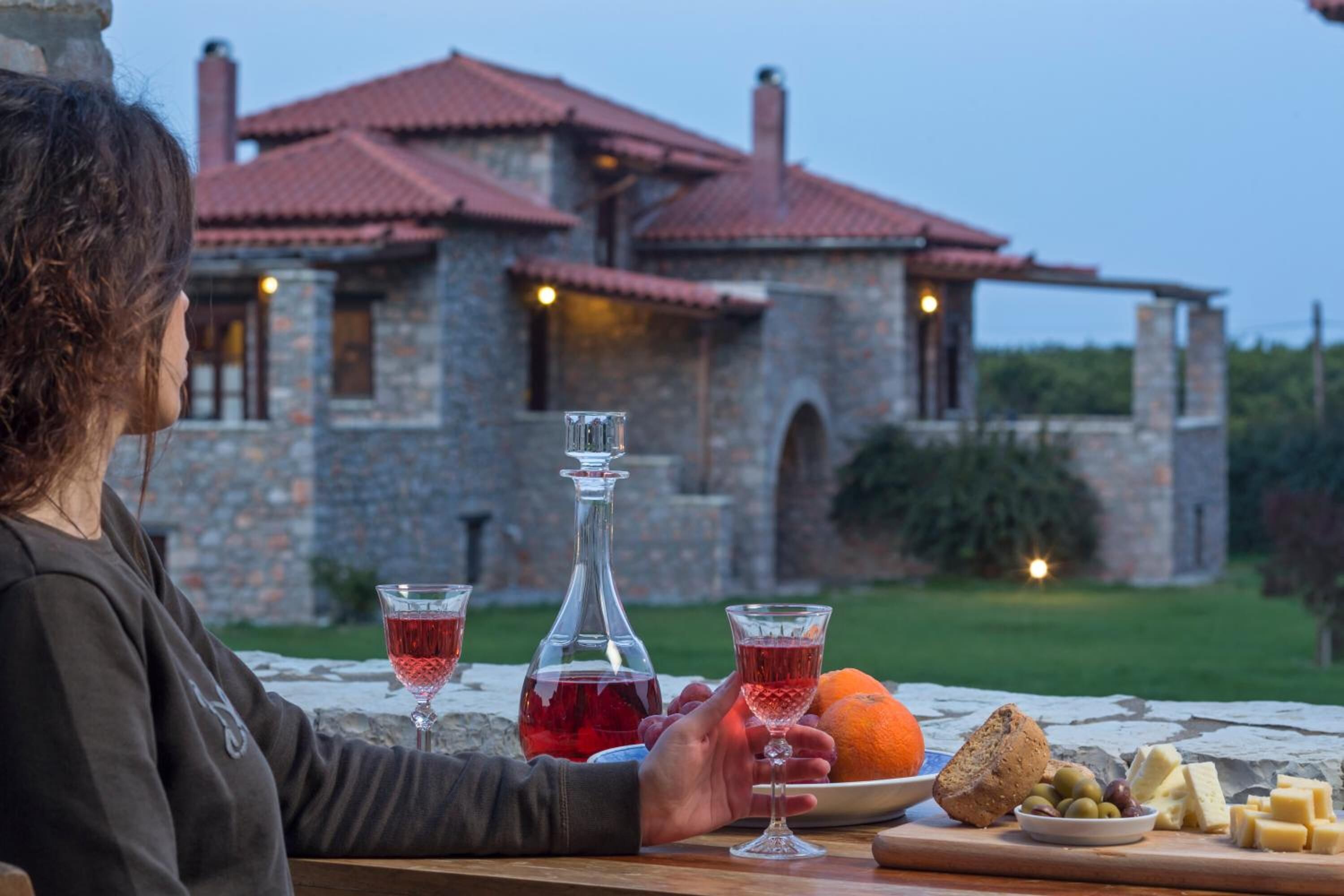 Une personne est assise à une table devant une bouteille de vin rouge, deux verres de vin rouge, une orange, des olives, du fromage et du pain. En arrière-plan, on aperçoit un bâtiment en pierre avec un toit rouge.