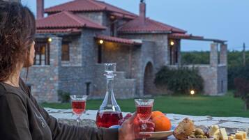 Une personne est assise à une table devant une bouteille de vin rouge, deux verres de vin rouge, une orange, des olives, du fromage et du pain. En arrière-plan, on aperçoit un bâtiment en pierre avec un toit rouge.