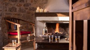 A rustic room with a stone wall, a wooden chair with a red cushion, a wooden table with a bottle, glasses, and a bowl of nuts, and a fireplace with lit candles. - Agroktima (South Kynouria)