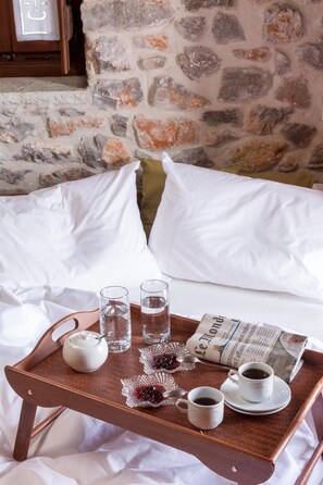 Una bandeja de madera con tetera, vasos, tazas y un periódico sobre una cama con almohadas blancas y una pared de piedra al fondo.