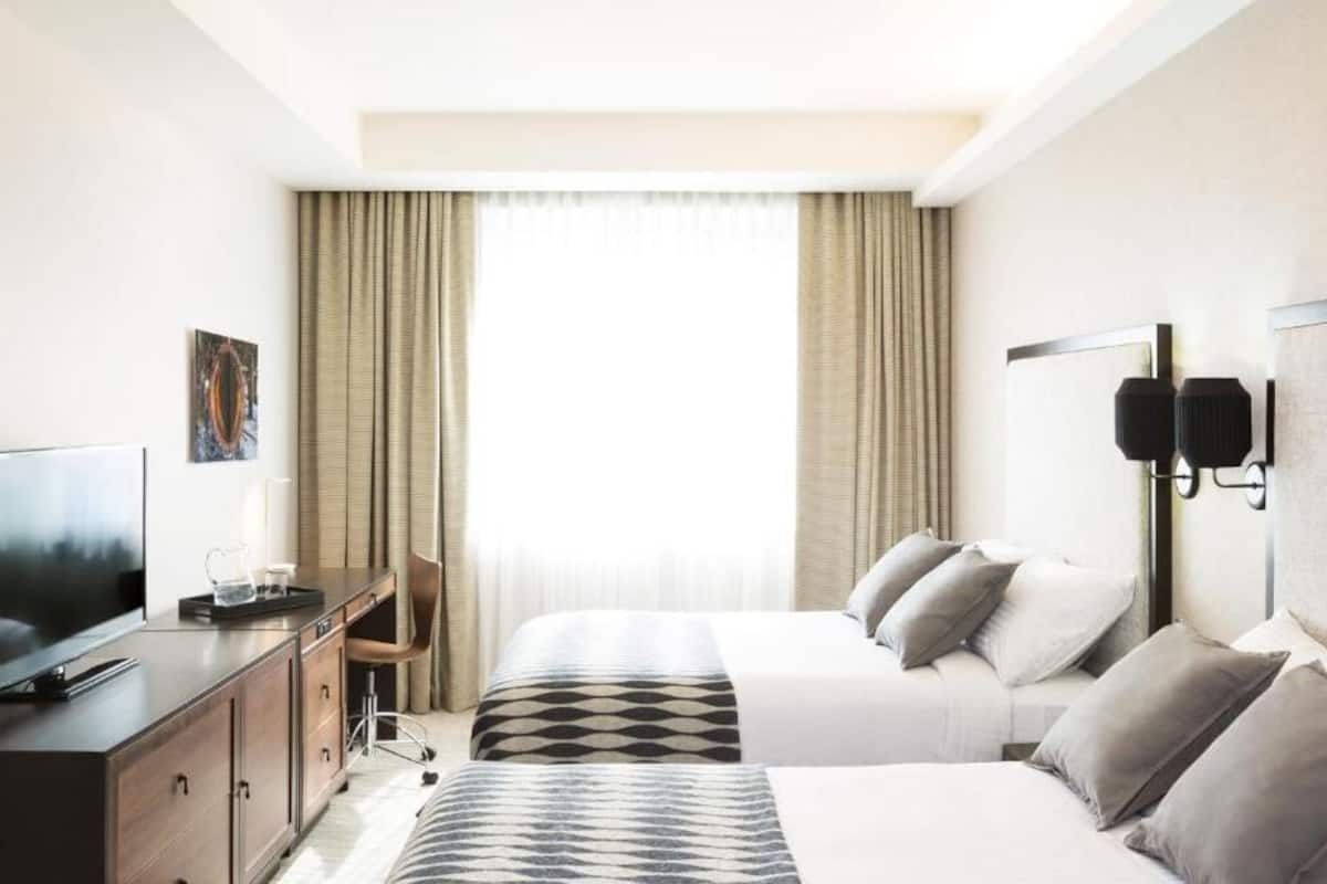 A hotel room with two beds, a desk with a TV, a chair, and a large window with curtains.
