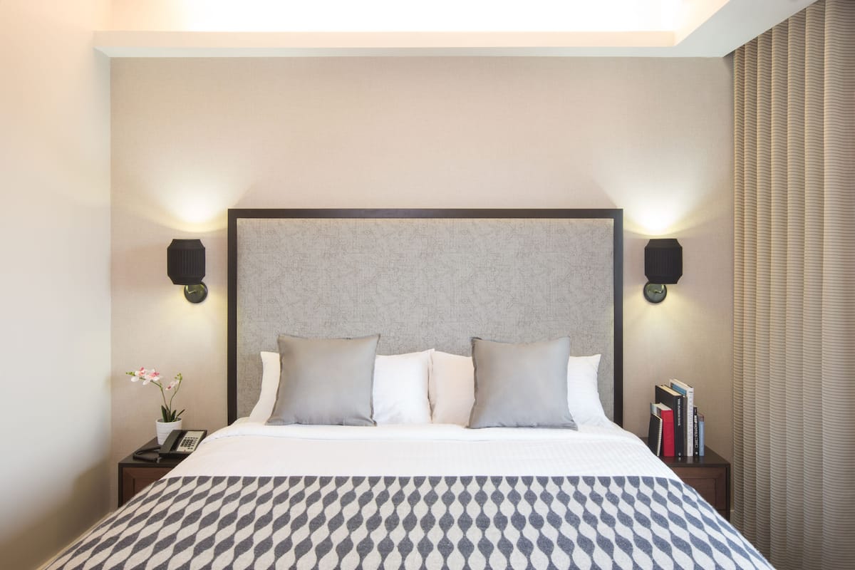 A neatly made bed with a patterned bedspread, two bedside tables with lamps, a clock, a potted plant, and a stack of books on one of the tables.