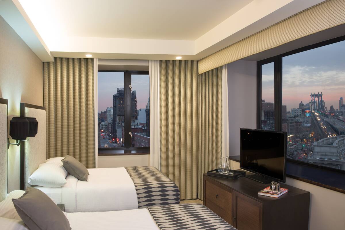 A hotel room with a bed, a TV, a desk, and a large window with a city view.