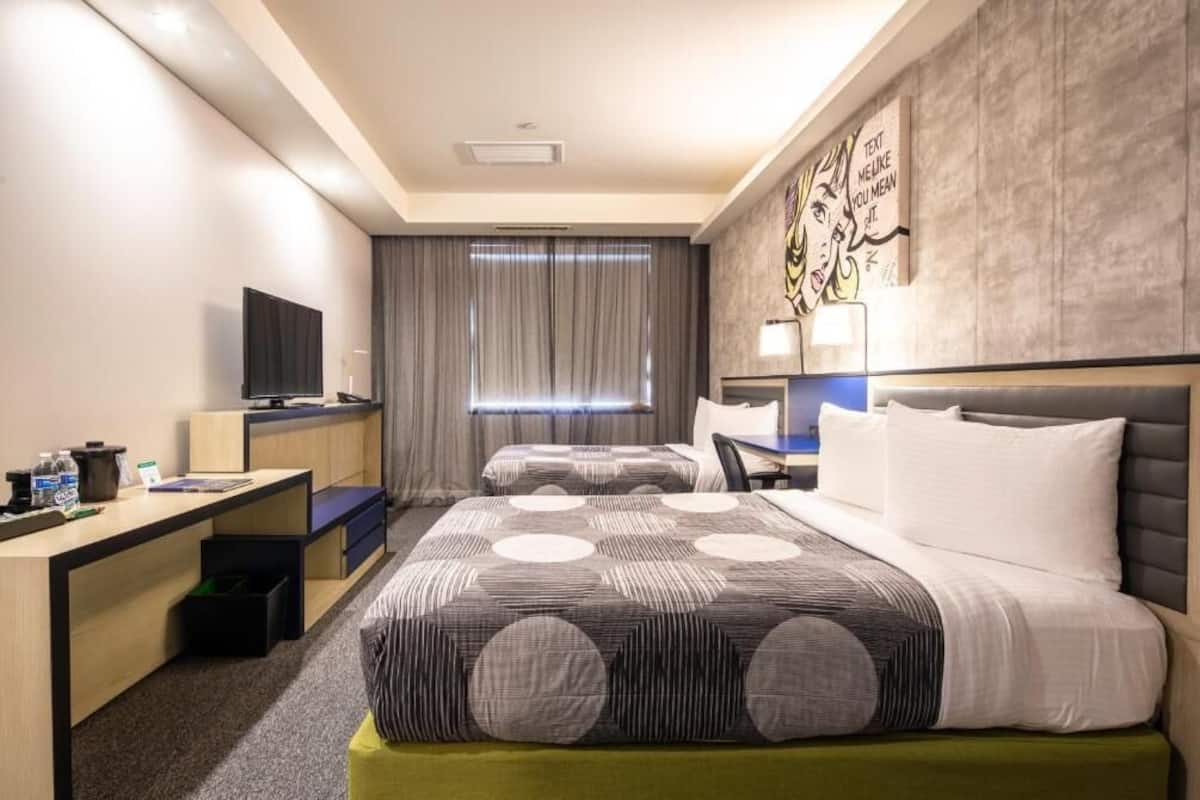 A hotel room with two beds, a desk, a television, and a large window with curtains.