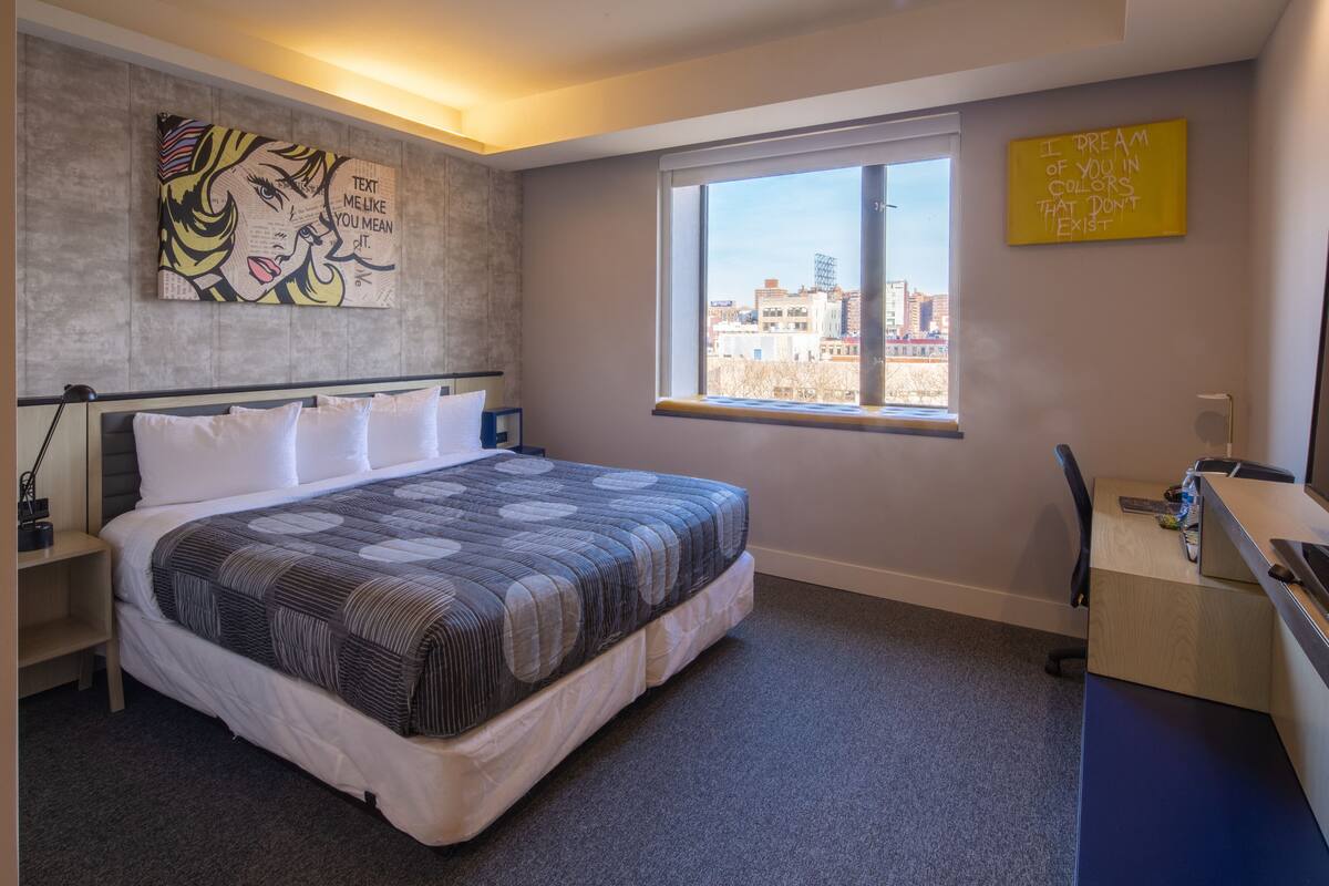 A modern bedroom with a large bed, a desk, and a window with a city view.