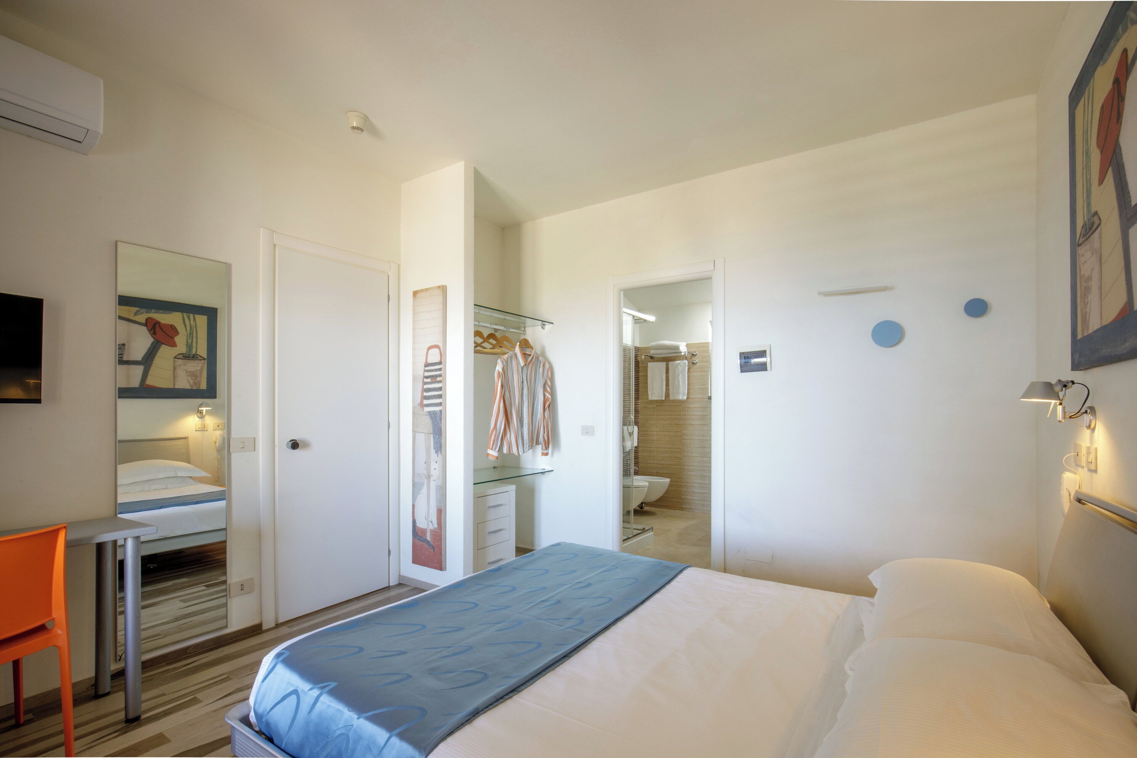 Habitación de hotel con cama, escritorio, silla, espejo, baño visible a través de una puerta abierta y un aplique de pared.