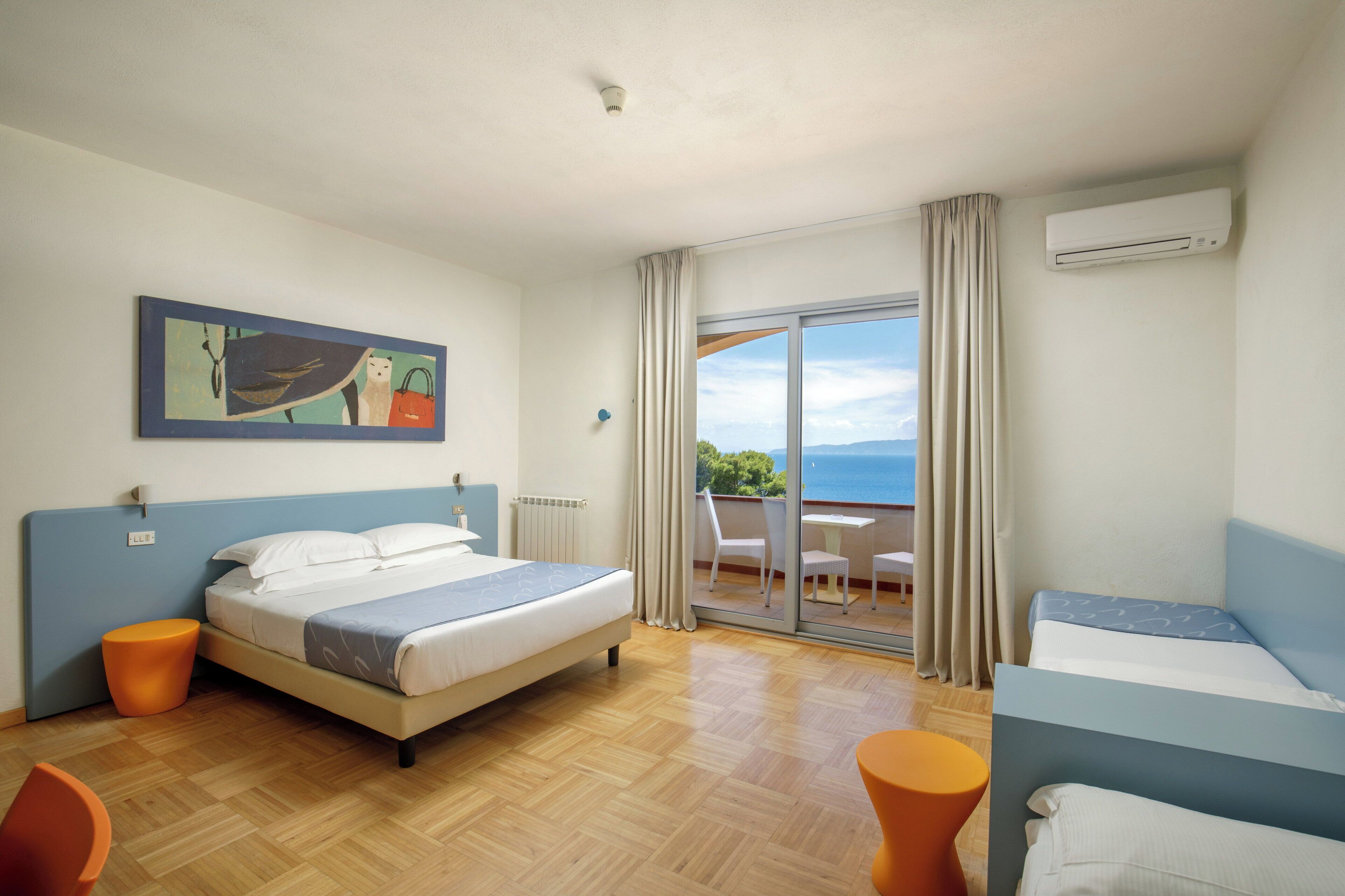 A hotel room with a bed, a small table, a chair, and a view of the sea through a sliding glass door.