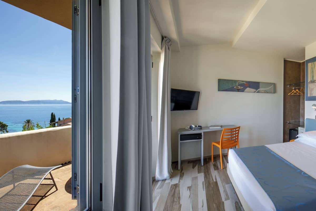 A hotel room with a bed, desk, chair, TV, and a balcony with a view of the sea.