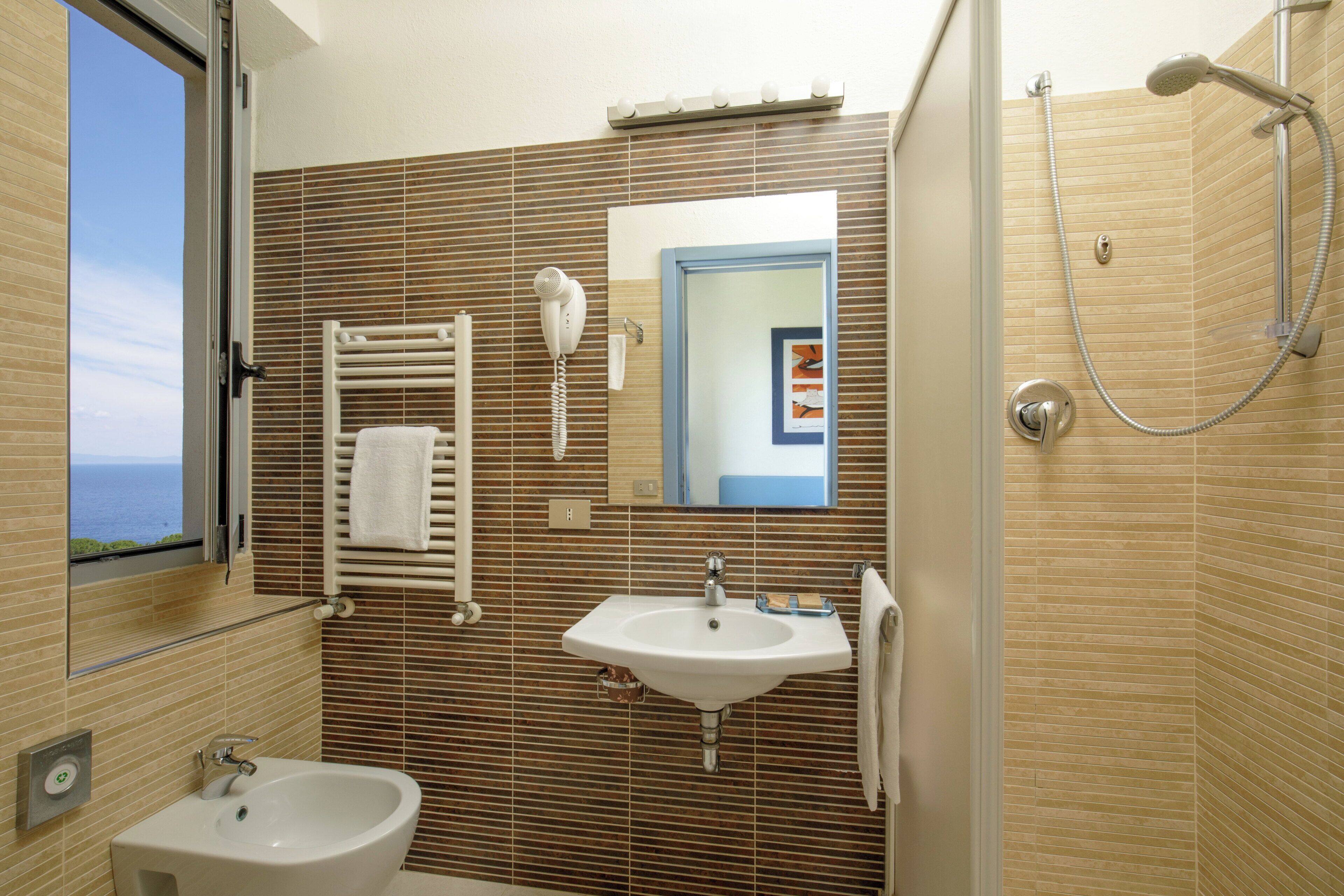 Une salle de bain dotée d’une grande fenêtre, d’un lavabo, d’un bidet, d’un porte-serviettes, d’un miroir et d’une douche.