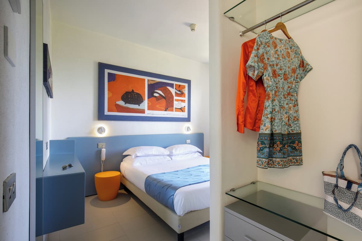 A modern hotel room with a bed, a bedside table, a glass-top desk, and a wall-mounted artwork.