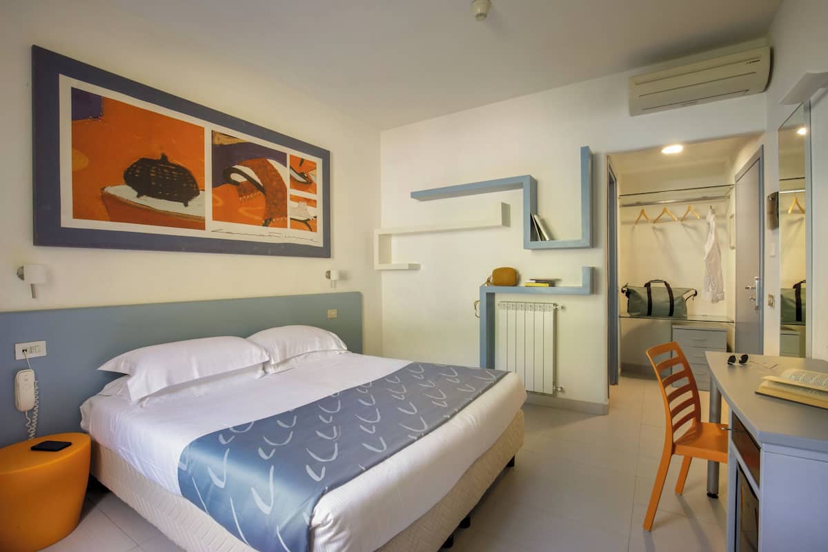 A hotel room with a bed, a desk, a chair, a bathroom with a mirror, and a wall-mounted air conditioner.