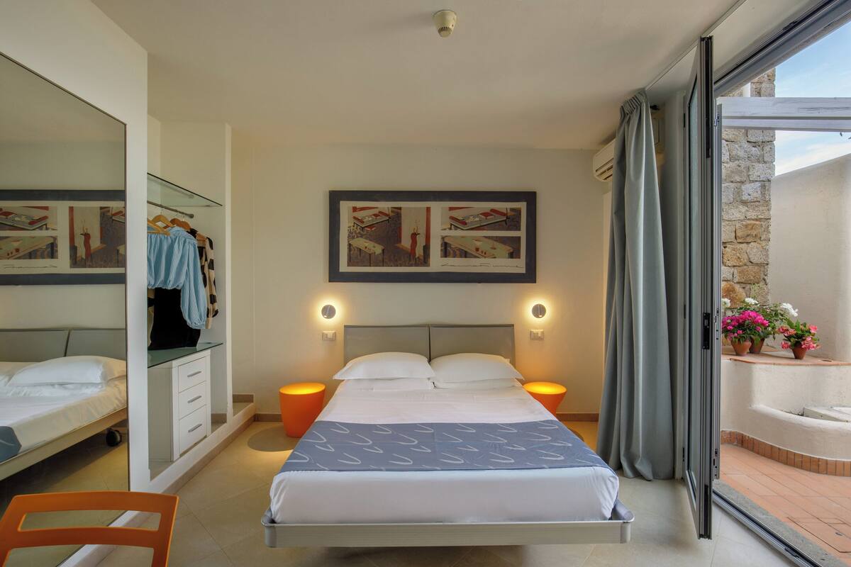 A bedroom with a bed, bedside tables, a mirror, a wardrobe, and a balcony with flowers.