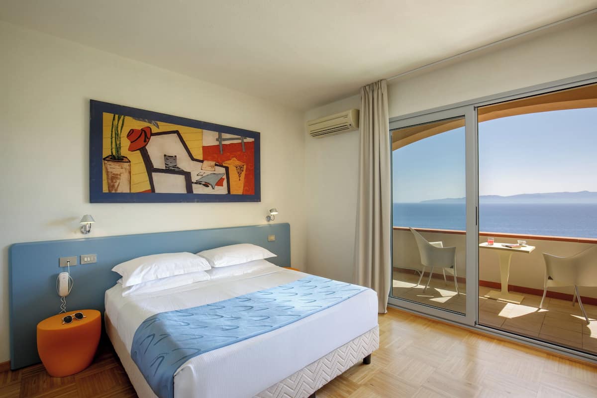 A hotel room with a bed, a painting on the wall, a phone, and a view of the sea through a balcony.