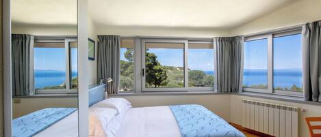 A bedroom with two beds, large windows, a view of the sea, and a wooden floor.