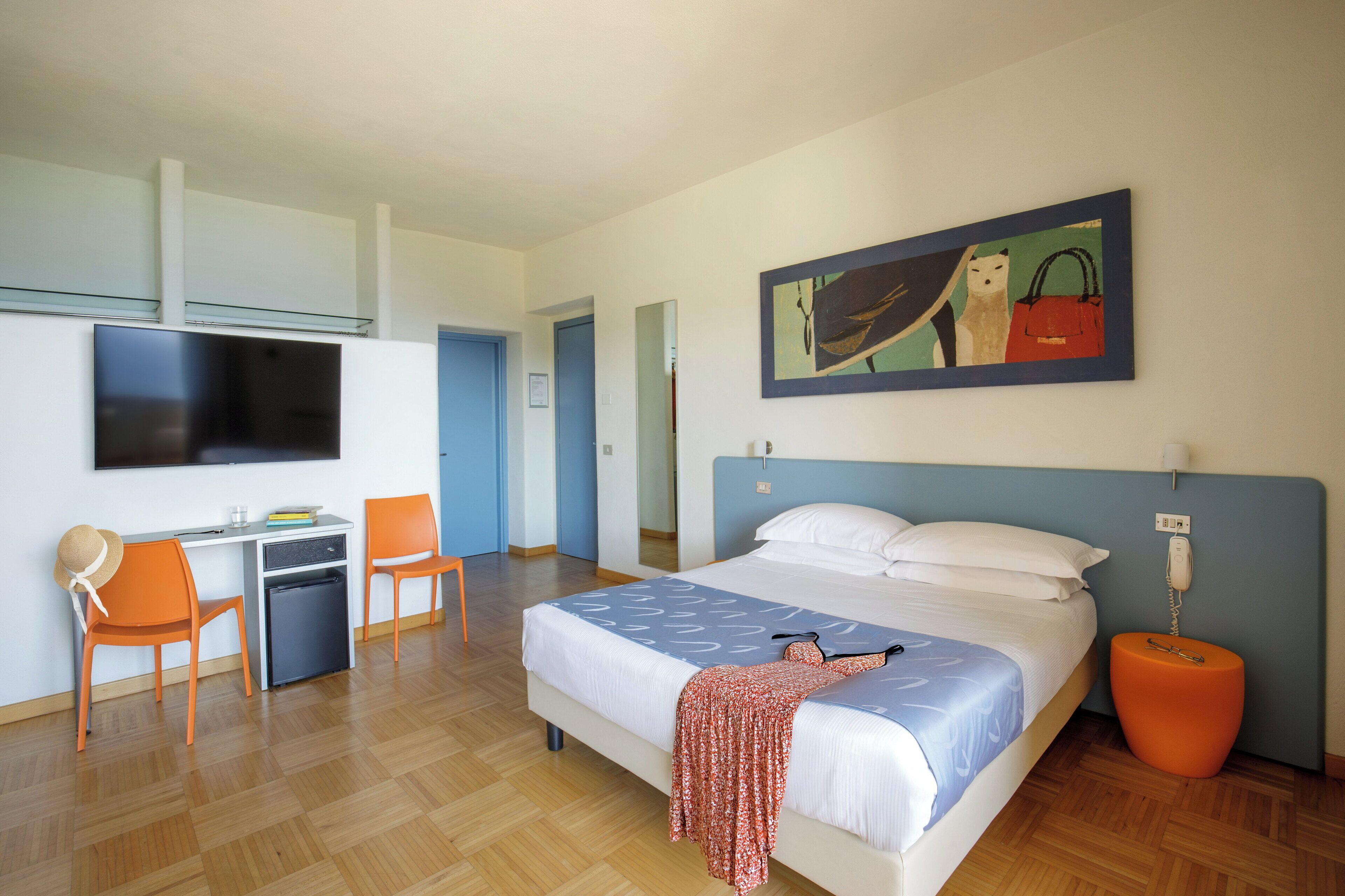 A hotel room with a bed, desk, chairs, a television, and a painting on the wall.