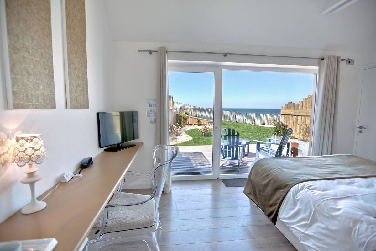 A hotel room with a bed, a desk with a TV, a lamp, and a view of the sea through a sliding glass door.