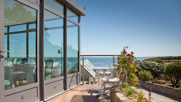 Um edifício moderno com grandes janelas de vidro, um deck de madeira, área de estar externa e vista para o mar.