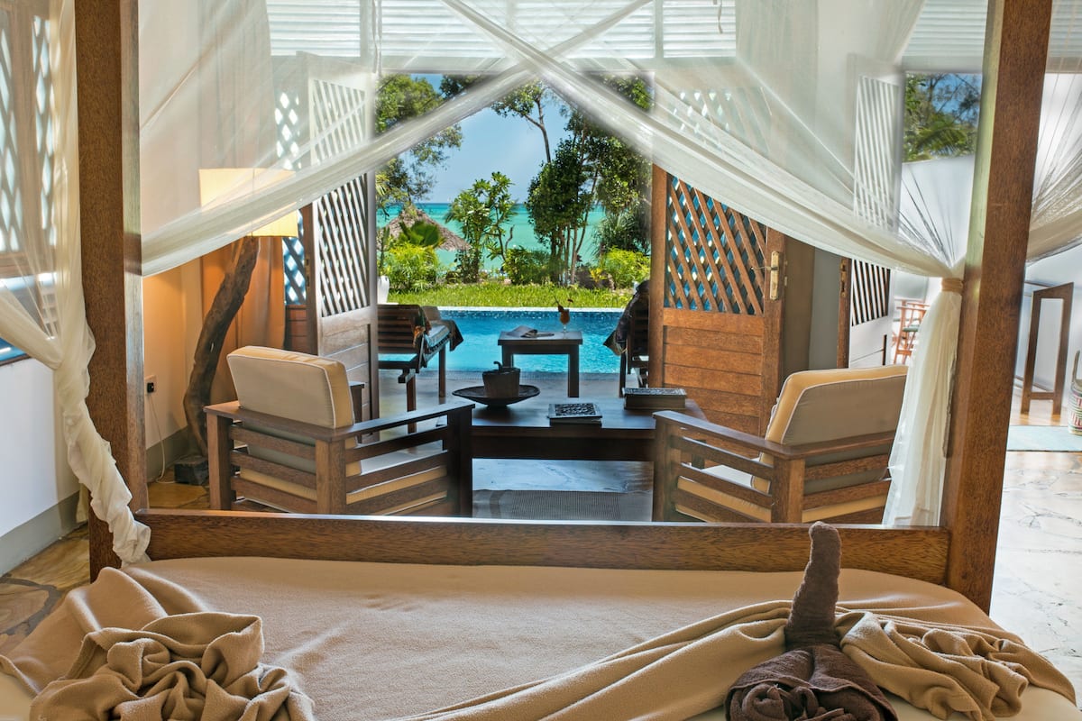 Habitación con vistas a una piscina y paisaje tropical, con una mesa y sillas de madera, una mesita con una lámpara y un espejo grande que refleja la vista.