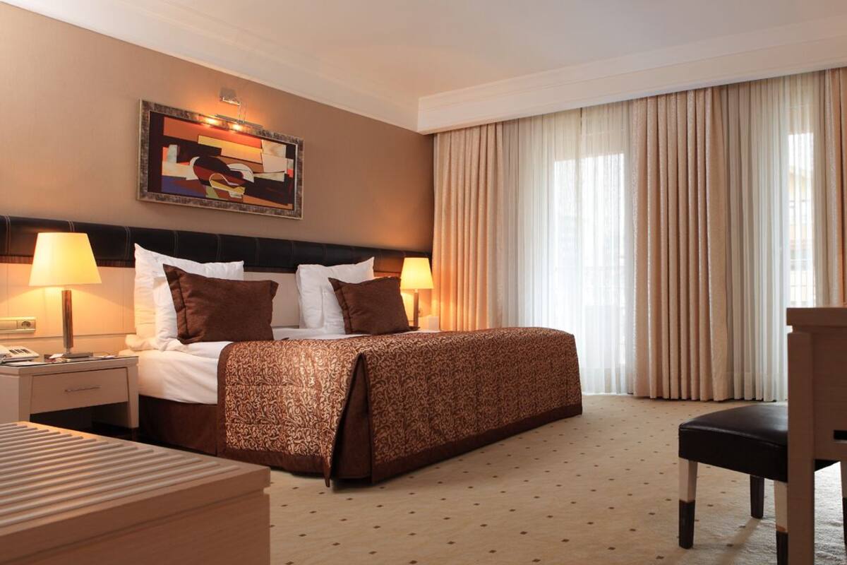 Habitación de hotel con cama, lámparas de noche, escritorio, silla, ventana grande con cortinas y un cuadro en la pared.