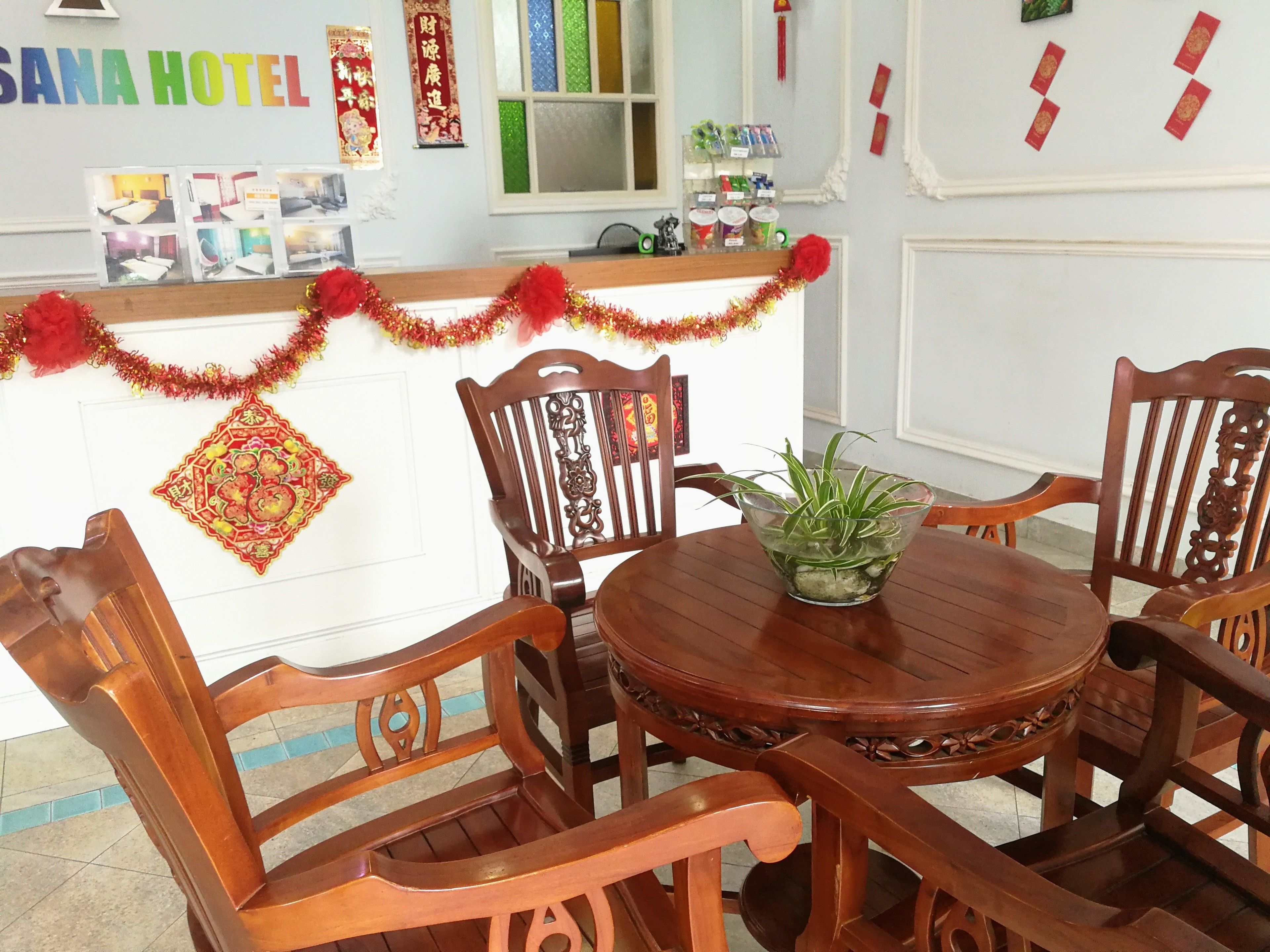 A wooden dining table with chairs, a decorative plant, and a wall with a sign reading "SANA HOTEL".