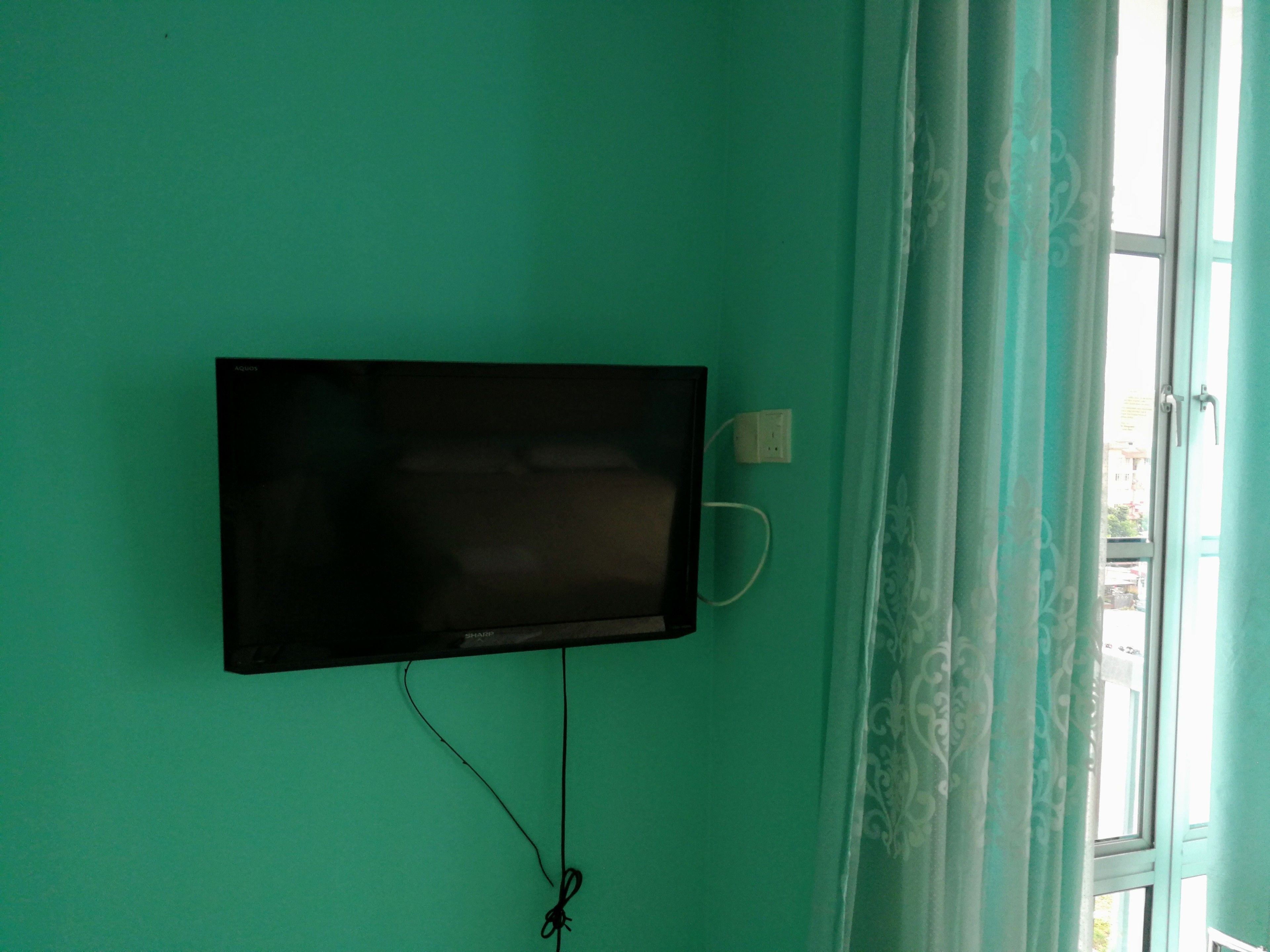 Quarto com televisão na parede verde-água, tomada e janela com cortinas.