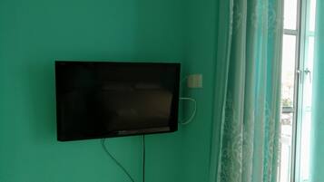Quarto com televisão na parede verde-água, tomada e janela com cortinas.