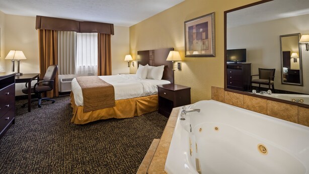 A hotel room with a bed, a desk, a chair, a desk lamp, a nightstand, a lamp, a large mirror, a TV, a bathroom with a bathtub, and a carpeted floor. - Best Western Inn (Elyria)