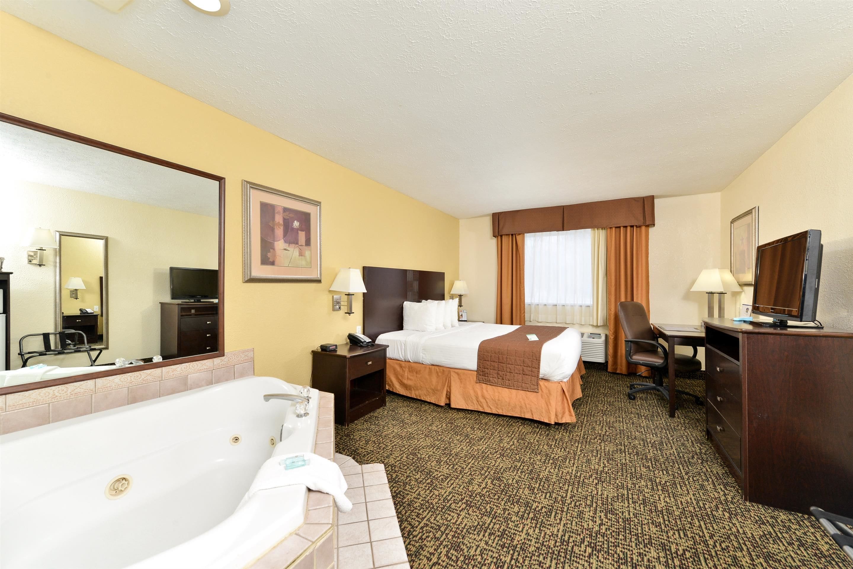 Quarto de hotel com cama grande, escrivaninha, cadeira, luminária de mesa, televisão, espelho, banheiro com banheira e janela com cortinas.