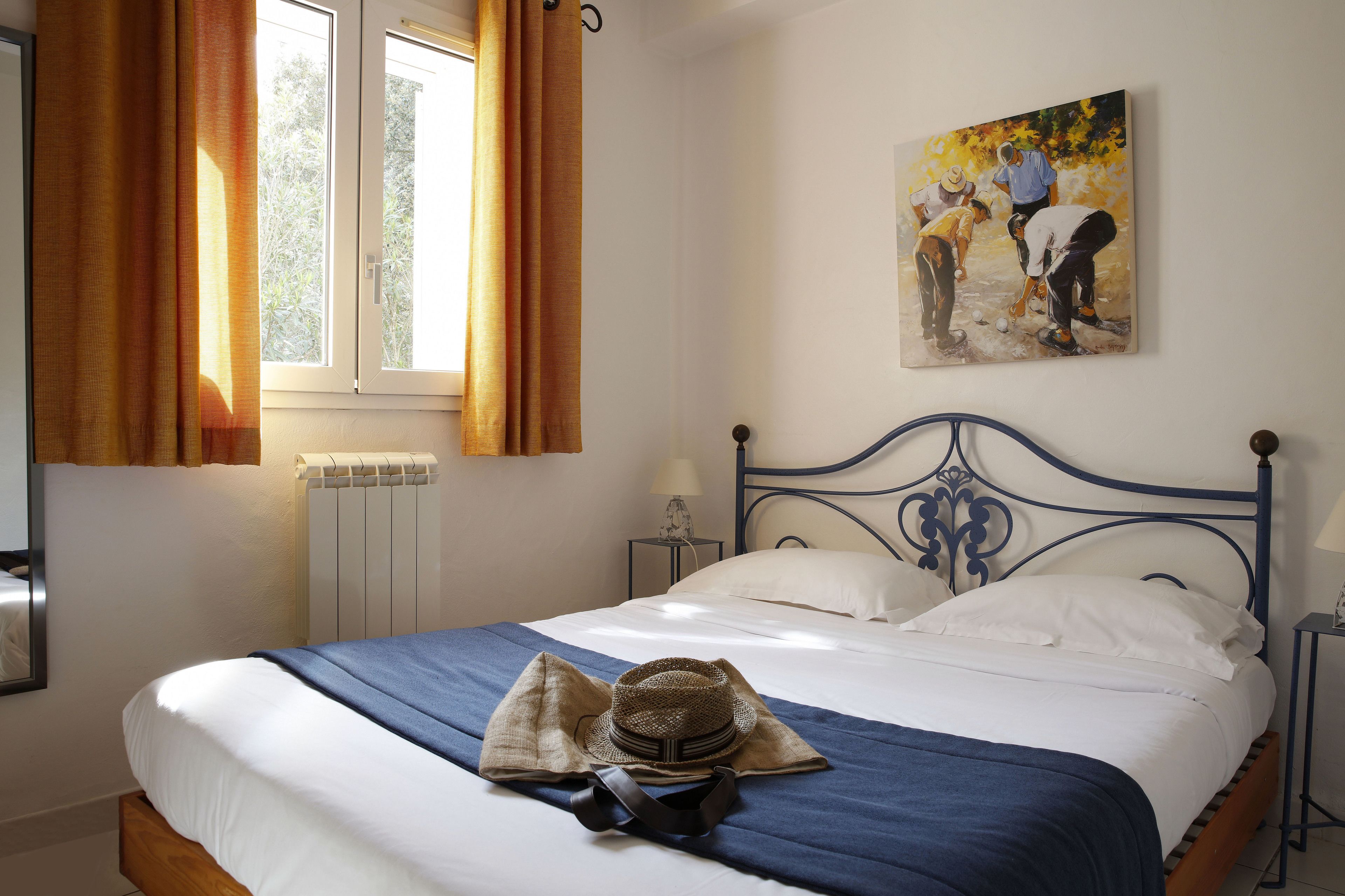 A bedroom with a bed, a radiator, a window with curtains, a painting on the wall, and a nightstand with a lamp.