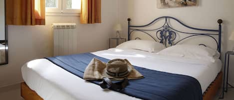 A bedroom with a bed, a radiator, a window with curtains, a painting on the wall, and a nightstand with a lamp.