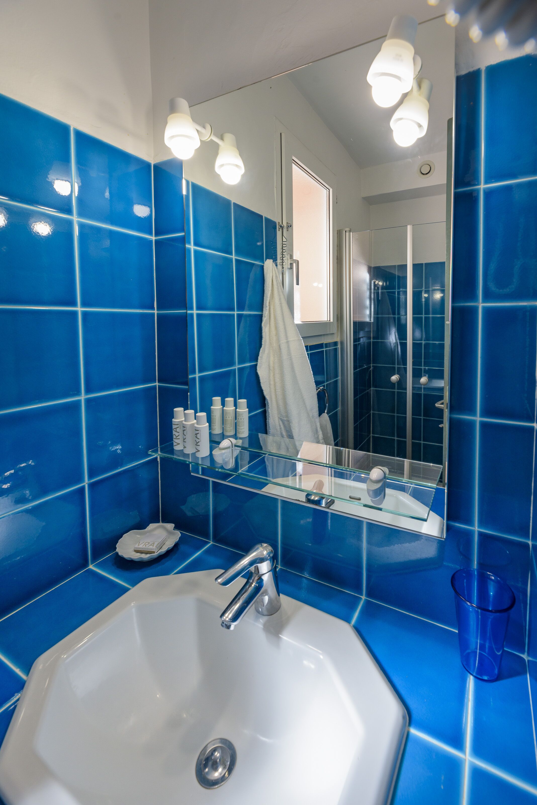 Une salle de bain avec des carreaux bleus, un lavabo blanc, une étagère en verre où sont rangés les articles de toilette et un miroir.