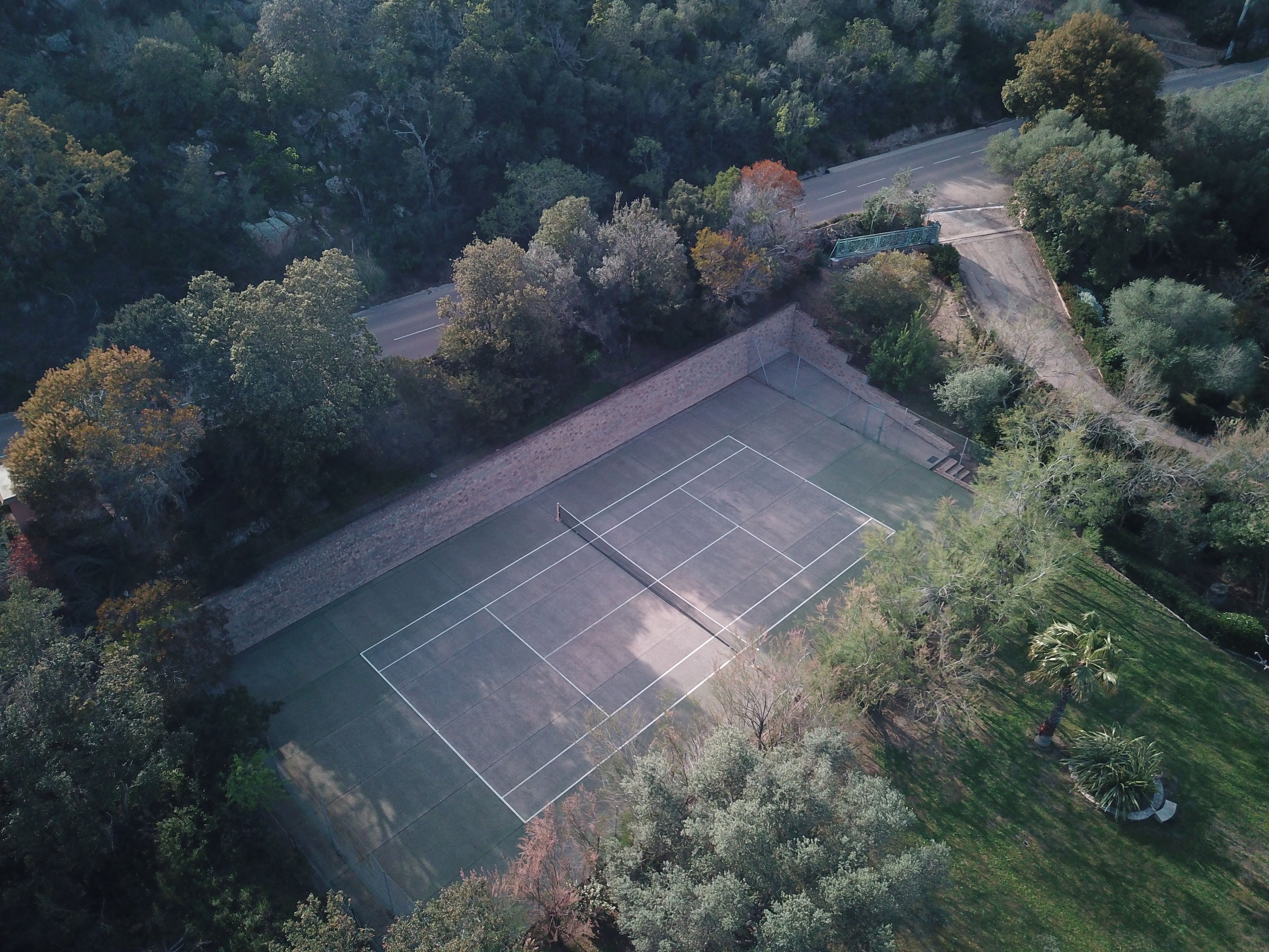 Une vue aérienne d’un court de tennis entouré d’une forêt dense et bordé d’une route.