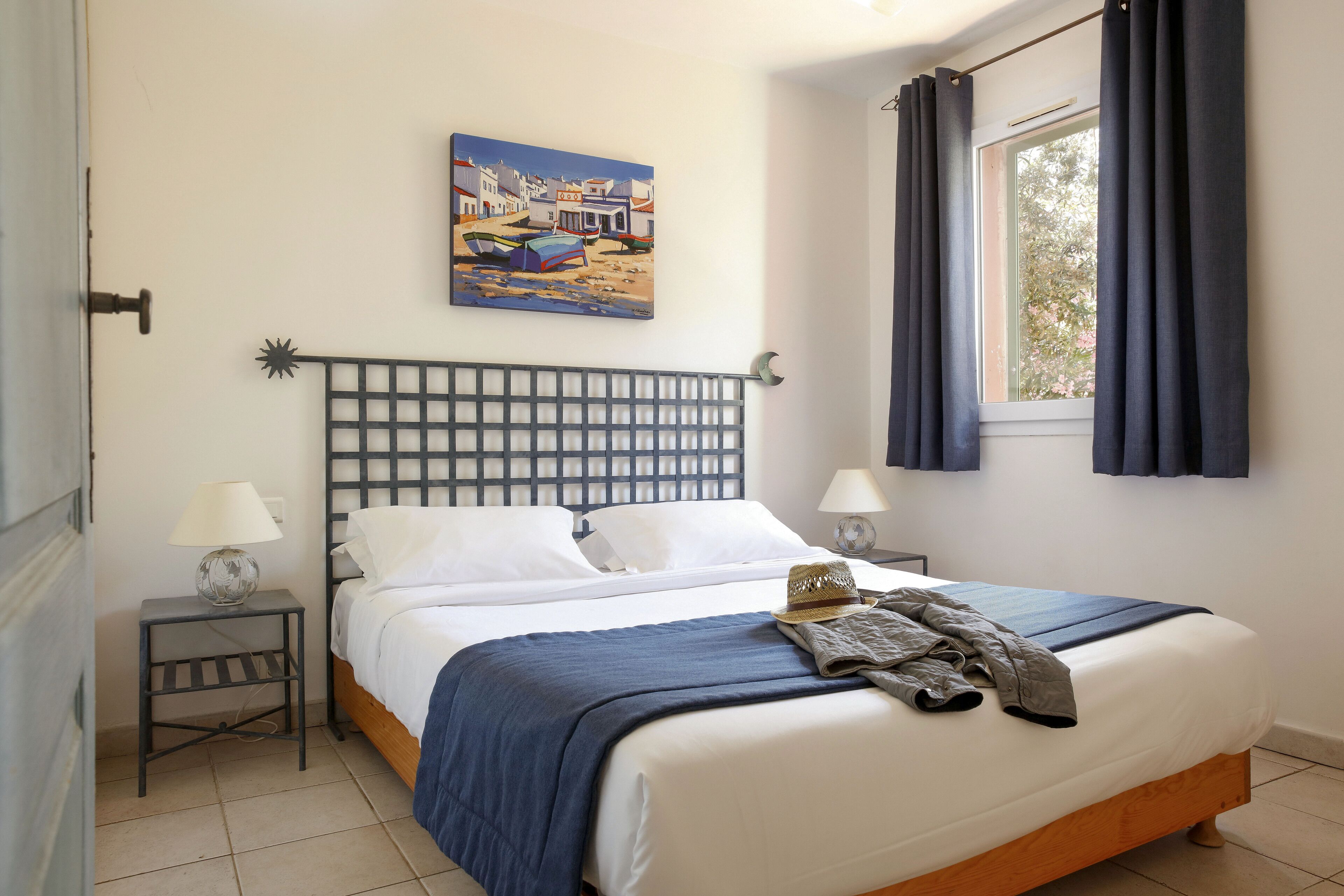 A neatly made bed with a blue blanket, two bedside lamps, a painting of a coastal scene, and a window with curtains.