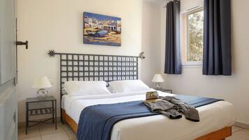 A neatly made bed with a blue blanket, two bedside lamps, a painting of a coastal scene, and a window with curtains.
