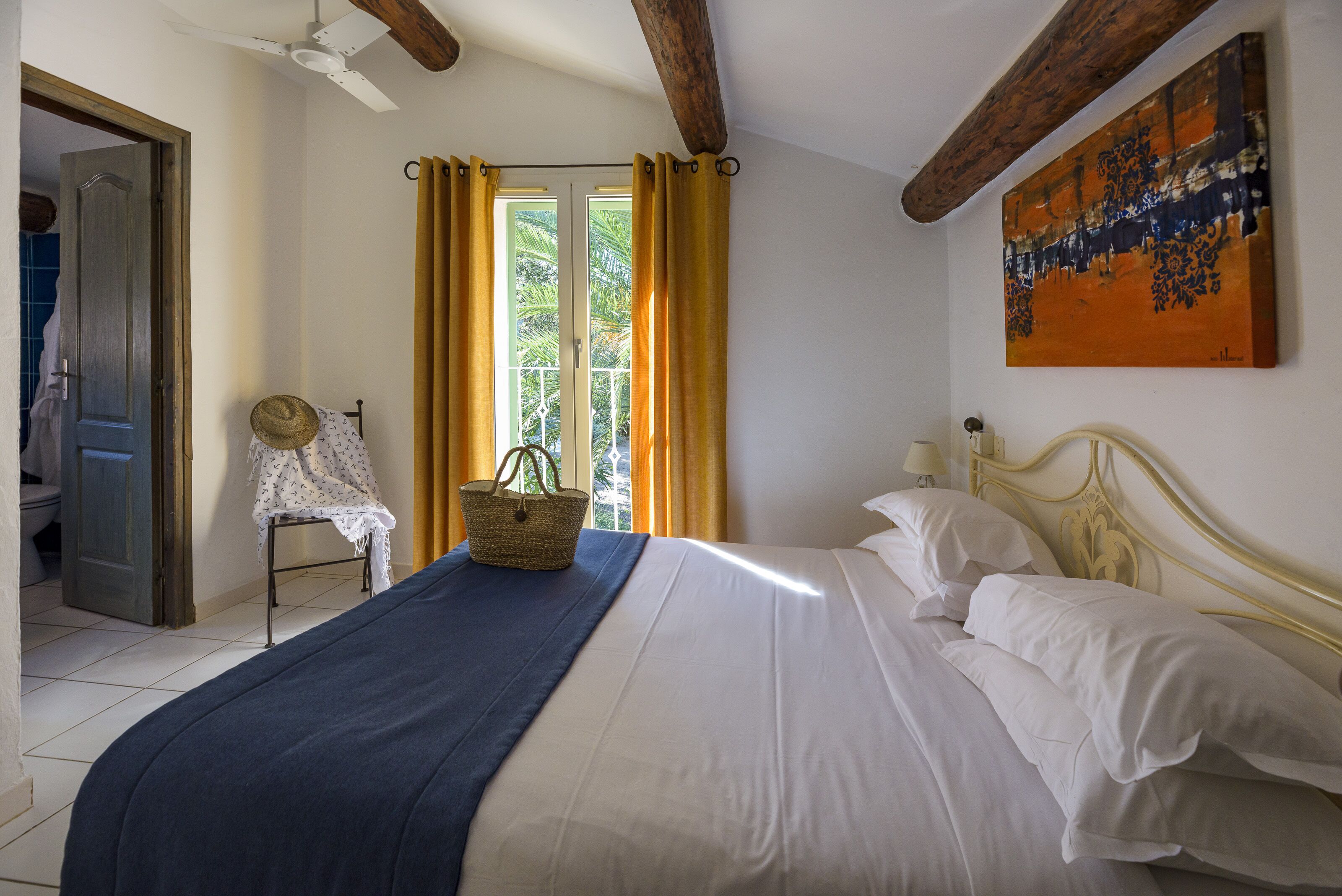 A bedroom with a bed, a window with curtains, a painting on the wall, and a door leading to another room.