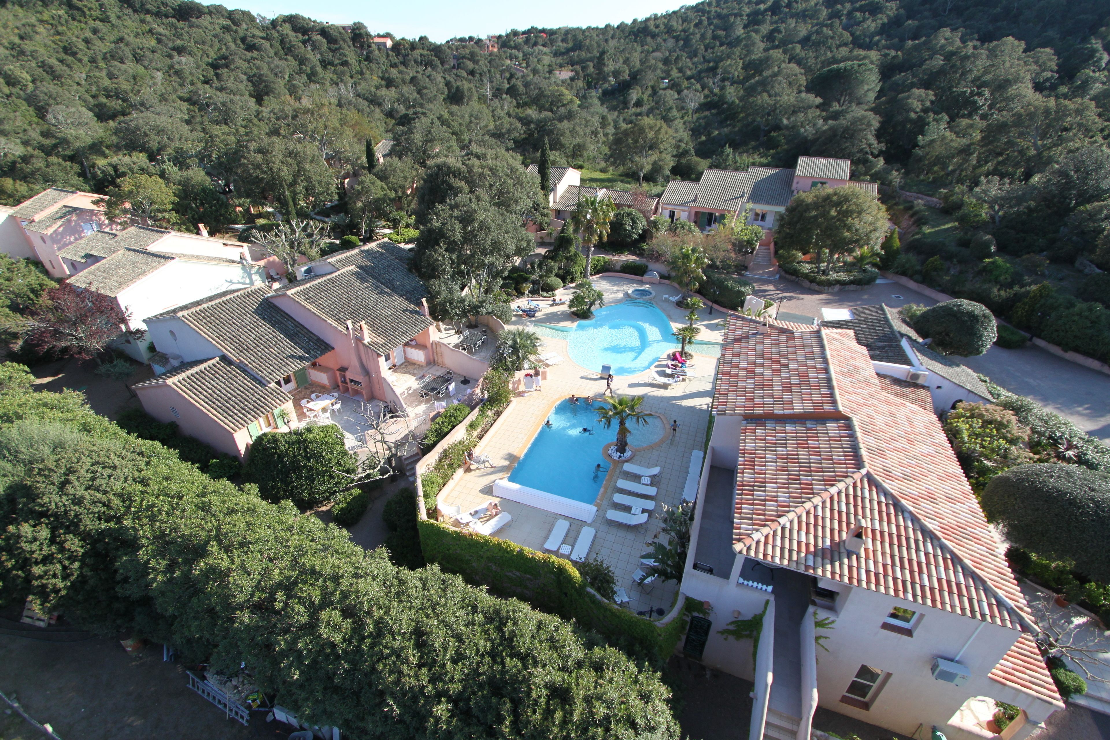 An aerial view of a resort with a swimming pool surrounded by lounge chairs and palm trees, set against a backdrop of lush greenery and terraced buildings.