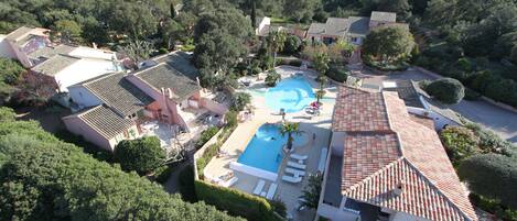 An aerial view of a resort with a swimming pool surrounded by lounge chairs and palm trees, set against a backdrop of lush greenery and terraced buildings.