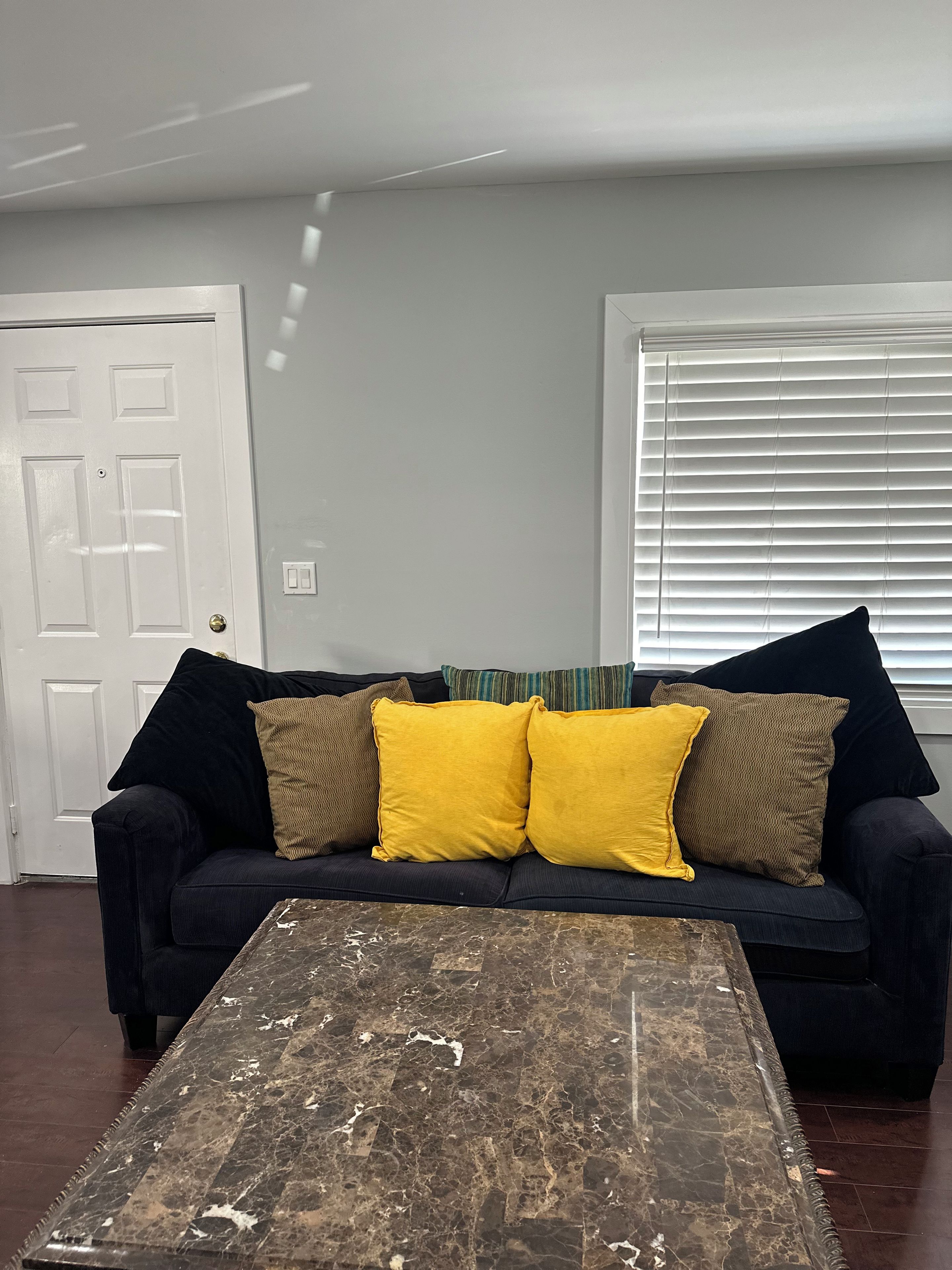 A living room with a dark-colored sofa, two yellow cushions, a wooden coffee table with a distressed finish, a white door, and a window with blinds.