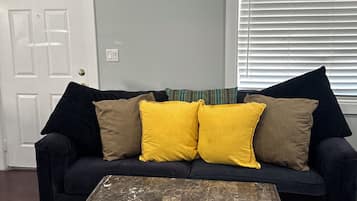 A living room with a dark-colored sofa, two yellow cushions, a wooden coffee table with a distressed finish, a white door, and a window with blinds.