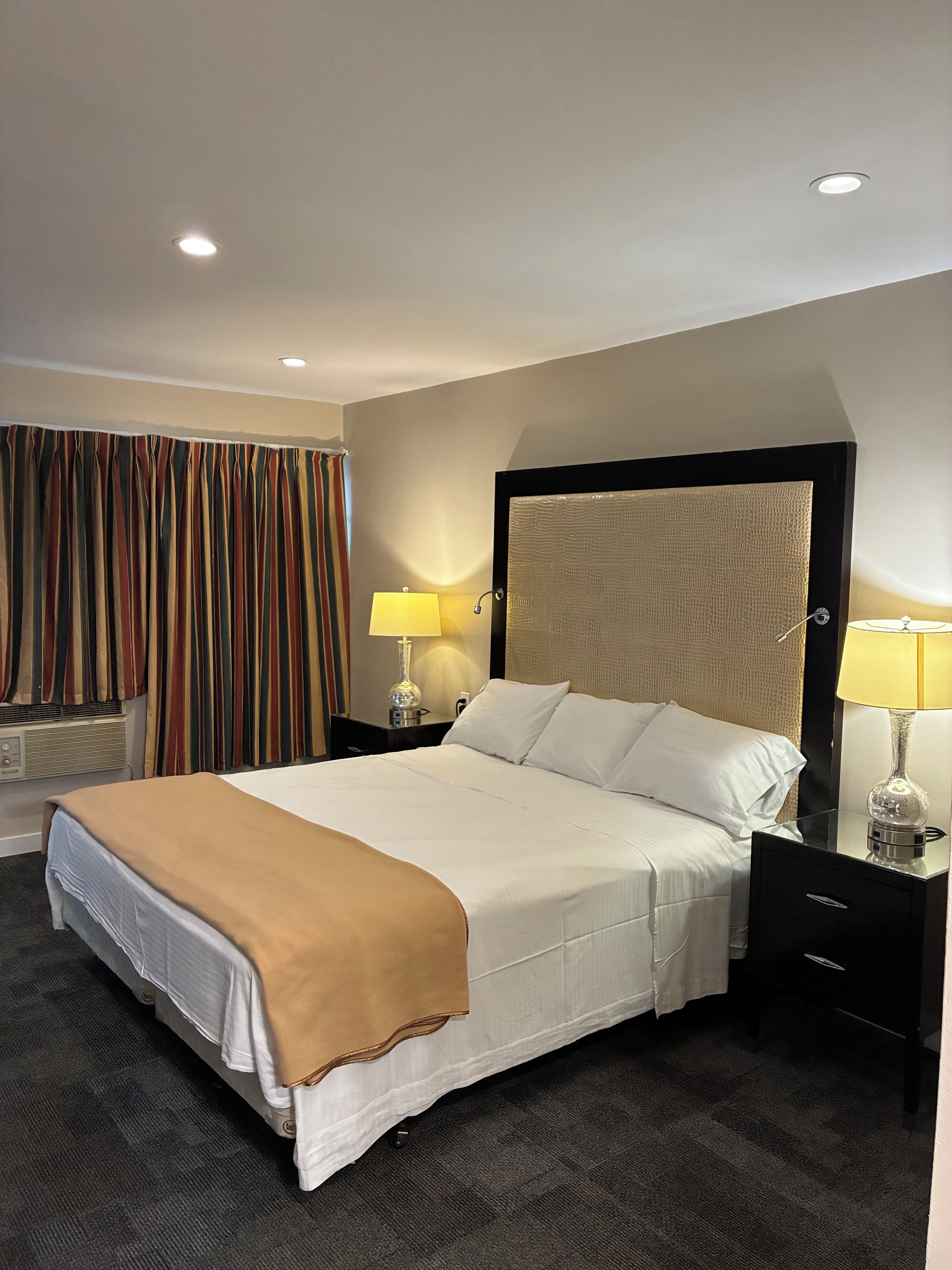 Una habitación de hotel con una cama grande, dos mesitas de noche, un cabecero y una ventana con cortinas.