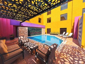 Un área de piscina con sillones de descanso, un muro de ladrillo y un edificio amarillo al fondo.