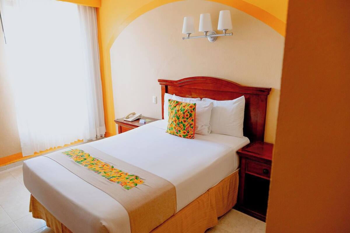 Habitación de hotel con cama, mesita de noche, teléfono e iluminación en la pared.