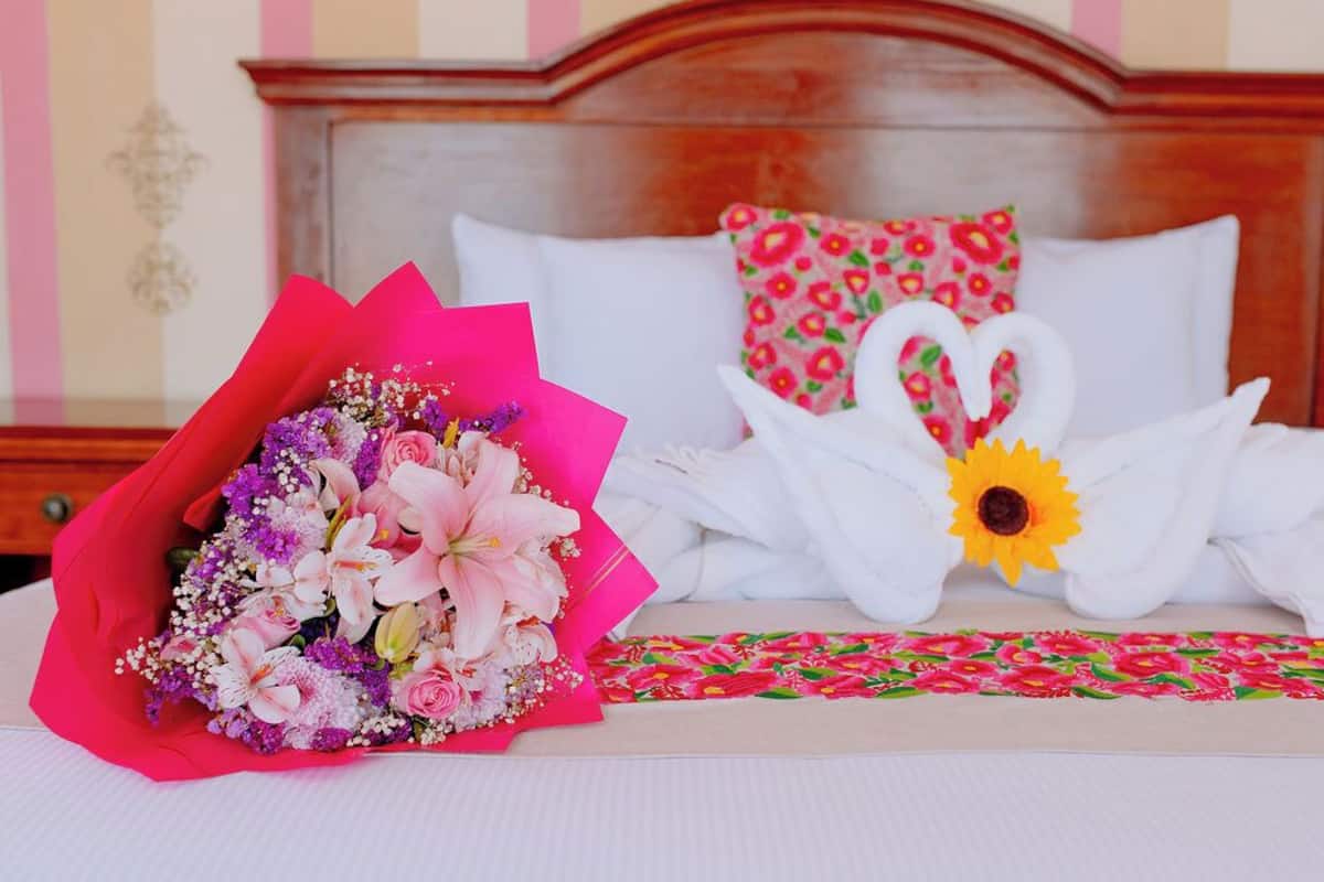 Una cama con almohadas blancas y una almohada con estampado floral, un ramo de flores y dos adornos con forma de cisne hechos de tela blanca.